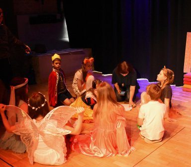 Grupa dzieci w kostiumach teatralnych siedzi na scenie podczas próby lub spektaklu. W centrum trwa rozmowa między uczestnikami przedstawienia, a dekoracje nawiązują do baśniowej scenerii.