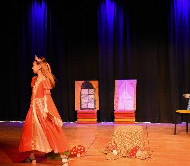Dziewczynka w różowej sukni i koronie stoi na scenie podczas występu teatralnego. W tle widoczne są proste dekoracje przedstawiające okna oraz elementy scenografii.