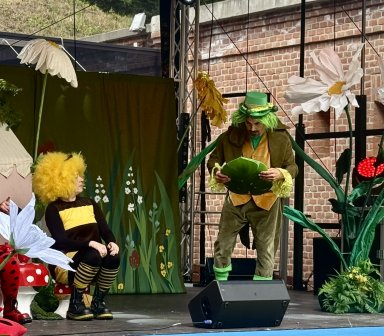 Scena spektaklu „Na pomoc ginącej przyrodzie” z udziałem kolorowych postaci oraz dekoracji przedstawiających łąkę i kwiaty.