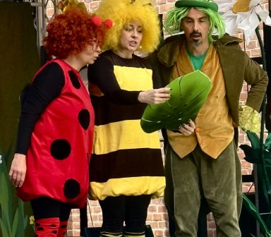 Na scenie plenerowej troje animatorów przebranych za owada, pszczołę i leśną postać odgrywa edukacyjny spektakl dla dzieci. W tle widoczna jest dekoracja przedstawiająca kwiaty i elementy przyrody.