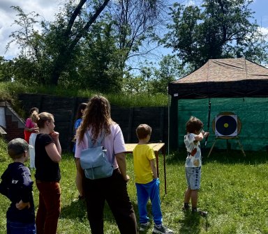 Dzieci i dorośli uczestniczą w plenerowych zajęciach łuczniczych na terenie Fortu Borek. Jedno z dzieci oddaje strzał do tarczy ustawionej na trawie.