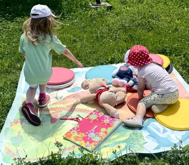 Najmłodsi uczestnicy pikniku bawią się na kolorowej macie edukacyjnej rozłożonej na trawie. Obok znajdują się zabawki i pomoce do zabawy sensorycznej.