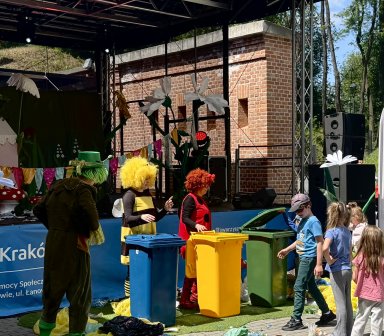 Animatorzy w barwnych kostiumach prowadzą ekologiczne warsztaty i zabawy związane z segregacją odpadów. Dzieci uczestniczą w zadaniach przy kolorowych pojemnikach do recyklingu.