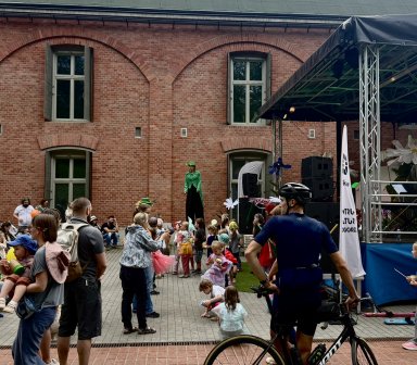 Dzieci wraz z rodzicami oglądają plenerowy spektakl na dziedzińcu Fortu Borek. Na scenie występują animatorzy na szczudłach i w kolorowych kostiumach.