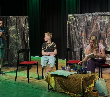 Kilkoro młodych aktorów odgrywa scenę przy stoliku nakrytym ozdobnym obrusem. Na scenie ustawiono krzesła i elementy dekoracji przedstawiające wnętrze magicznej chaty.