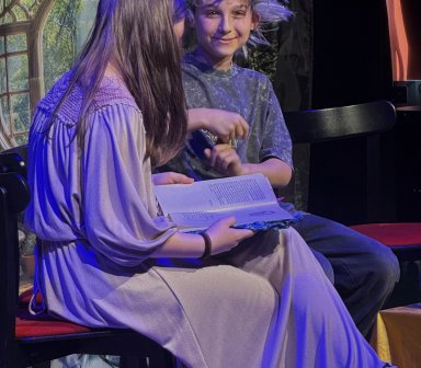 Dwójka uczestników oraz postać w fantazyjnej peruce siedzą przy stoliku podczas jednej ze scen spektaklu. Kolorowe światła sceniczne podkreślają baśniowy charakter przedstawienia.