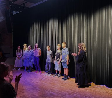Finał spektaklu „Jak oswoić czarownicę?” – wszyscy uczestnicy warsztatów teatralnych stoją na scenie podczas ukłonów, a publiczność nagradza ich występ brawami.