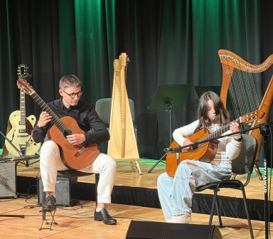 Instruktor i uczestniczka zajęć siedzą na scenie z gitarami klasycznymi i wspólnie wykonują utwór. W tle stoją harfa oraz instrumenty przygotowane do koncertu.