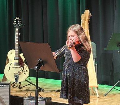 Młoda skrzypaczka w ciemnej błyszczącej sukience gra na skrzypcach przed pulpitem nutowym. W tle widoczne są gitara, wzmacniacze i harfa.