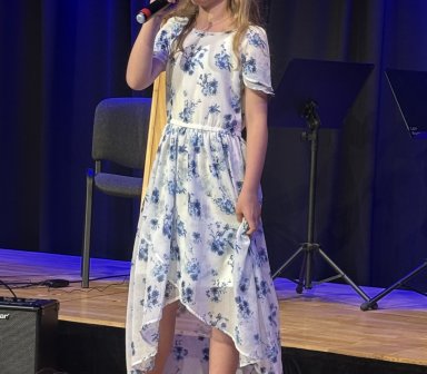 Młoda wokalistka w białej sukience w niebieskie kwiaty śpiewa do mikrofonu na scenie Fortu Borek. Za nią stoją krzesło i pulpit muzyczny.