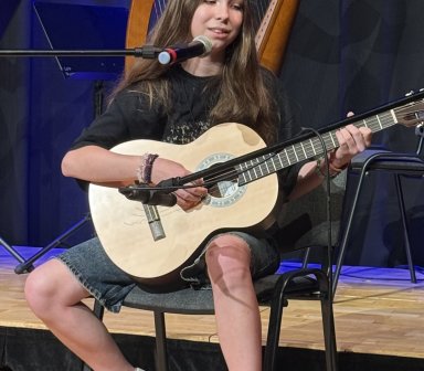 Uczestniczka koncertu siedzi z gitarą klasyczną i śpiewa do mikrofonu. Za nią widoczna jest harfa oraz ciemne kurtyny oświetlone niebieskim światłem.