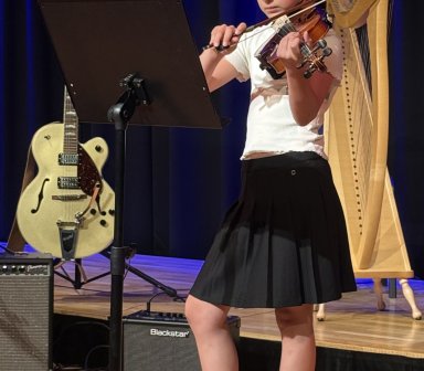 Młoda skrzypaczka gra na skrzypcach przed pulpitem z nutami. W tle znajdują się gitara, harfa oraz wzmacniacze ustawione na scenie.