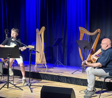 Młody uczestnik koncertu śpiewa i gra na gitarze elektrycznej, występując wspólnie z instruktorem grającym na gitarze akustycznej. Na scenie Fortu Borek widoczne są również harfy, wzmacniacze oraz pulpity muzyczne, a całość oświetla niebieskie światło.