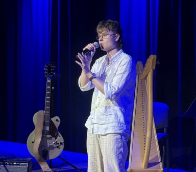 Młody wokalista w jasnym stroju śpiewa do mikrofonu na scenie Fortu Borek. W tle znajdują się harfa, gitara i elementy wyposażenia muzycznego.
