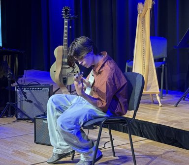 Uczestnik zajęć siedzi na krześle i gra na gitarze elektrycznej podczas koncertu finałowego. W tle widoczne są fortepian, harfa oraz sprzęt nagłośnieniowy.