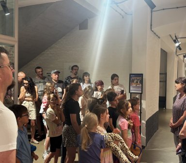 Uczestnicy wernisażu zgromadzeni przy wystawie prac plastycznych dzieci i młodzieży. Wśród publiczności znajdują się mali artyści wraz z rodzinami, a prowadzące prezentują ekspozycję zawieszoną na ceglanej ścianie galerii.